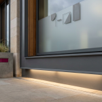 Safety First - Professional 16:9 hero banner. Ultra high resolution. A close-up, architectural detail shot of a modern, well-maintained commercial building entrance in the UK, possibly an educational institution or corporate office. The entrance features clear signage (no text, just abstract shapes) and a subtle, reassuring glow of the accent colour #737373 emanating from beneath a sturdy, well-secured door frame, symbolising security and compliance. The primary colour #E2087E is subtly integrated into some structural elements or a planted feature nearby. The overall mood is authoritative and trustworthy, with strong lines and a sense of permanence. Style: Architectural photography with a focus on clean design and subtle lighting. Avoid: Groups around holographic tables, generic tech aesthetics, text overlays, cold blue tones, stock photo clichés.