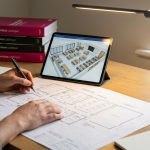 School Furniture Project - Professional 16:9 hero banner. Ultra high resolution. Close-up, slightly overhead shot of a School Business Manager's hands, in a contemporary school office setting, meticulously reviewing a detailed blueprint or architectural drawing for a classroom layout. One hand holds a pen, poised over the plans, while the other rests on a tablet displaying 3D rendering software. The scene is illuminated by a subtle, focused glow of #737373 light emanating from the tablet screen and a desk lamp, highlighting the intricate details of the plans. A background element of a stack of educational furniture catalogues in shades of #E2087E and #000000 is softly blurred, suggesting comprehensive planning. Style: Editorial photography, focusing on precision and strategic thinking. Avoid: Groups around holographic tables, generic tech aesthetics, text overlays, cold blue tones, stock photo clichés.