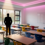 School Furniture Replacement - Professional 16:9 hero banner. Ultra high resolution. A confident, authoritative school business manager stands silhouetted against a softly blurred, modern school classroom interior. The manager is reviewing a digital project plan displayed on a tablet, with key elements of the classroom (ergonomic chairs, flexible tables) subtly visible. Accent lighting in #737373 casts a subtle glow around the manager and the tablet, highlighting the strategic planning aspect. The background features hints of #E2087E and #000000 in the furniture or wall details. The overall mood is one of organised control and successful project execution. Style: Editorial photography, conveying trust and expertise. Avoid: Groups around holographic tables, generic tech aesthetics, text overlays, cold blue tones, stock photo clichés.