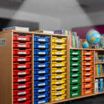 School Storage Solutions - Professional 16:9 hero banner. Ultra high resolution. A meticulously organised school storage shelf, filled with colour-coded trays and educational resources. A beam of light, accentuating the order and efficiency, glows in #737373 from above, highlighting a label with 'BS EN 1729' on a sturdy, well-constructed storage unit. The background subtly features the vibrant #E2087E and #000000 tones, suggesting a modern, well-equipped UK classroom environment. The focus is on the quality of the furniture and the clarity of organisation, symbolising expertise and compliance. Style: Editorial still life photography, with sharp focus and depth of field. Avoid: Groups of people, cluttered messy scenes, generic office furniture.