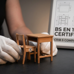 School Furniture Procurement - Professional 16:9 hero banner. Ultra high resolution. A close-up, focused shot of a pair of hands, likely belonging to a school business manager, carefully examining a miniature, perfectly compliant school desk and chair (BS EN 1729 certified, with a tiny 'Size Mark' visible). The furniture models are pristine, reflecting the importance of quality. In the background, subtly blurred, are technical specifications or a compliance certificate, with key text like 'BS EN 1729' and 'Crib 5' highlighted by a #737373 accent glow. The overall atmosphere is authoritative and precise, with a professional, documentary photography style. Hints of #E2087E and #000000 are incorporated into the background elements or as subtle reflections. Avoid: Groups around holographic tables, generic tech aesthetics, text overlays, cold blue tones, stock photo clichés.