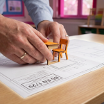 Ergonomics & Posture - Professional 16:9 hero banner. Ultra high resolution. A close-up, editorial style shot of a pair of adult hands, possibly a school business manager or architect, carefully aligning a small, colourful wooden ruler or scale model of a school table with a detailed blueprint or technical drawing that features the 'BS EN 1729' standard mark prominently. The scene is set on a clean, light-coloured desk surface, with a subtle glow of #737373 accentuating the edges of the blueprint and ruler. Hints of #E2087E can be seen in the background as a soft, out-of-focus educational setting. The overall mood is precise, authoritative, and focused on compliance. Style: Macro detail photography with a shallow depth of field. Avoid: Groups around holographic tables, generic tech aesthetics, text overlays, cold blue tones, stock photo clichés.