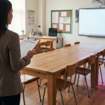 School Furniture Issues - Professional 16:9 hero banner. Ultra high resolution. A single, authoritative figure, a School Business Manager (SBM) or facilities professional, stands confidently in the foreground of a modern, well-maintained school classroom. Their back is partially to the viewer, looking towards a meticulously organised classroom with sturdy, compliant tables and chairs, each item appearing solid and in perfect condition. The SBM holds a clipboard with a checklist. A subtle, focused glow in #737373 emanates from key points on the furniture, highlighting its structural integrity and safety, casting a slight reflection of #E2087E on the polished floor. The overall atmosphere is one of proactive management and reliability, emphasising a safe learning environment. Style: Editorial documentary photography, with precise focus and a clean, professional aesthetic. Avoid: Groups around holographic tables, generic tech aesthetics, text overlays, cold blue tones, stock photo clichés.
