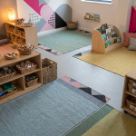 Stimulating EYFS Environments - Professional 16:9 hero banner. Ultra high resolution. An intricately designed, colourful EYFS classroom. The scene is viewed from a slightly elevated perspective, showcasing distinct 'learning zones' subtly defined by low-level, compliant furniture and different coloured floor rugs. One zone features natural 'loose parts' like wooden blocks, pinecones, and fabric scraps, bathed in a soft, warm glow of #737373 accent lighting. Another zone shows a cosy reading nook with cushions. Geometric patterns subtly integrated into the background, hinting at strategic planning and structured learning, using elements of #E2087E and #000000. The overall atmosphere is organised, inviting, and highly functional for young children. Style: Editorial photography with a focus on interior design and child-centric elements. Avoid: Groups around holographic tables, generic tech aesthetics, text overlays, cold blue tones, stock photo clichés.